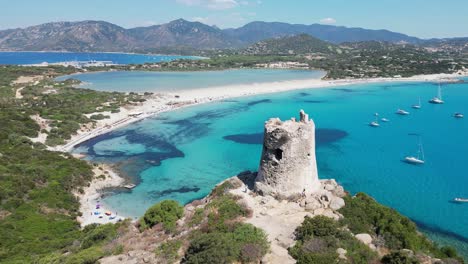 tourist people walk to porto giunco beach tower in villasimius, sardinia, italy - aerial 4k circling