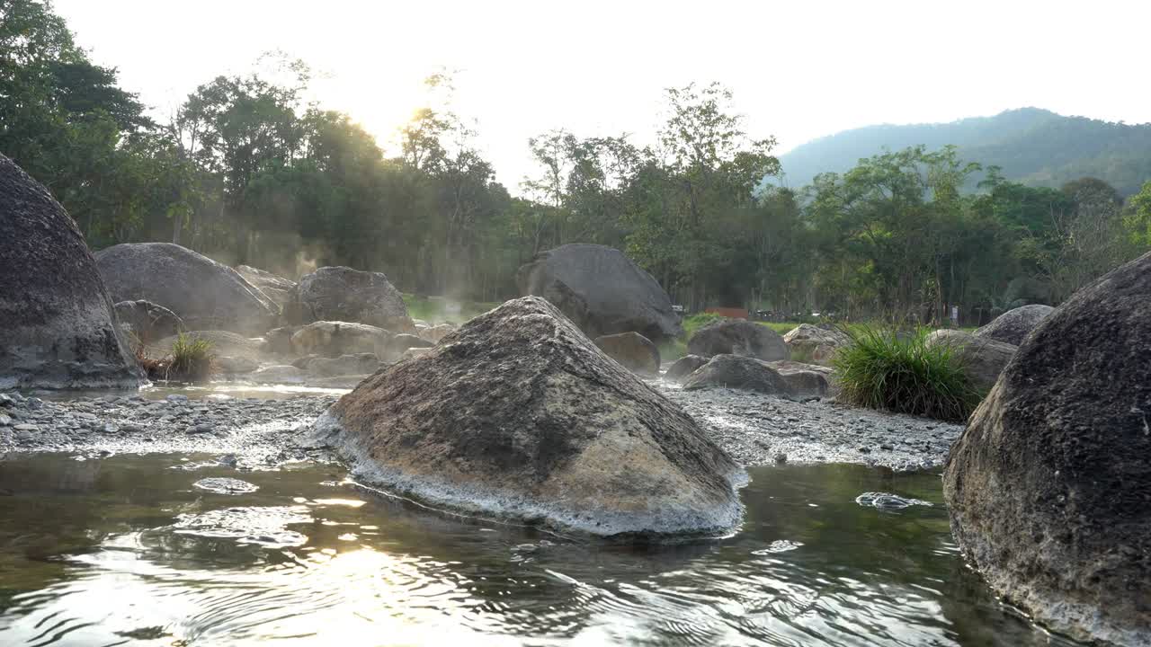4k disparo estático cambio de enfoque de aguas termales naturales fluyen a lo largo de las rocas bajo la luz del sol de la noche, el vapor flotando con árboles y hierba de fondo. parque nacional chaeson, lampang, tailandia.