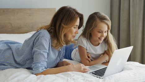 junge schöne mutter, die mit ihrer tochter auf dem bett liegt und morgens etwas auf dem laptop anschaut