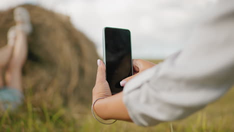 close-up hand view of lady in long sleeve taking photo of someone leg placed on parked hay, the background is slightly blurred, highlighting the focus on smartphone photography in rural scenery