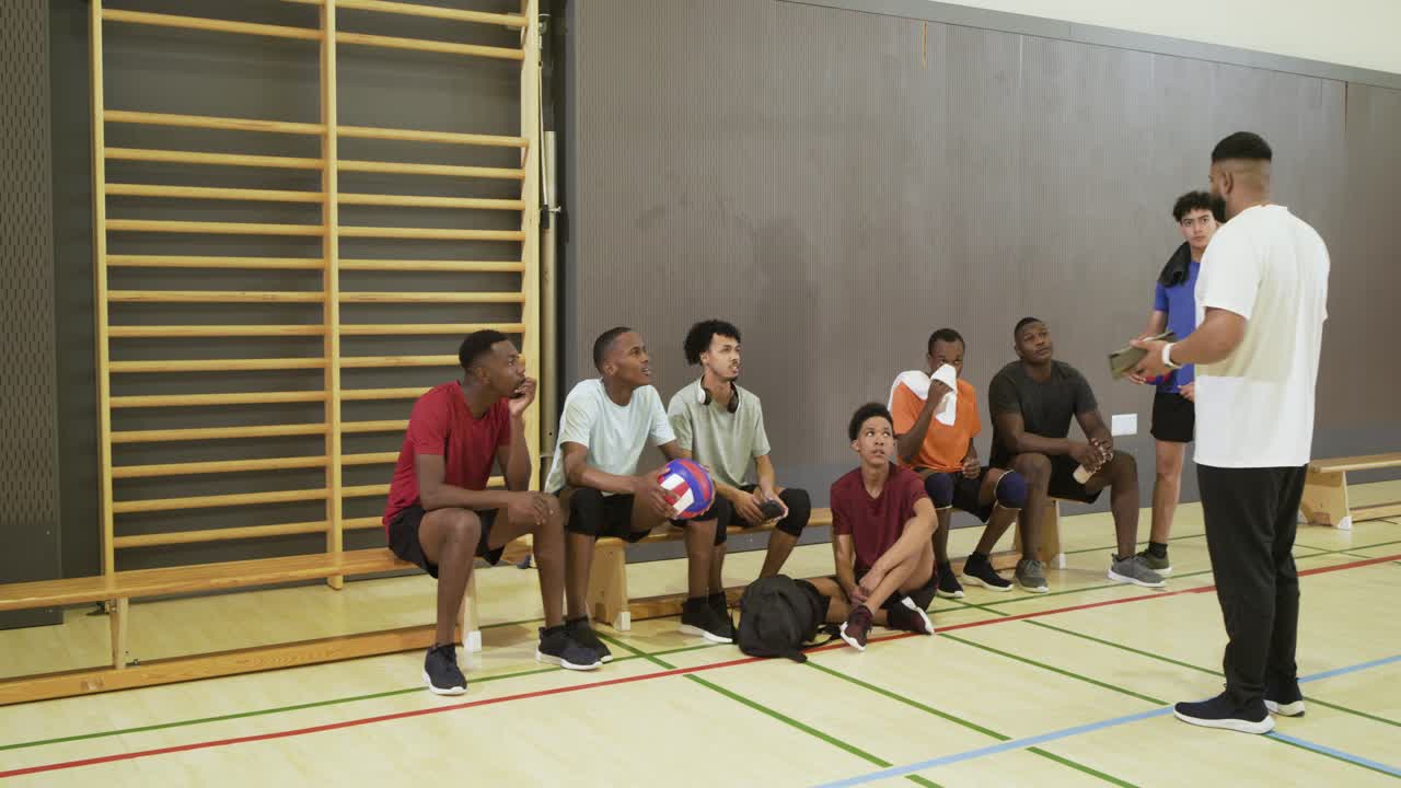 Vielfältiges männliches Volleyballteam mit Bällen; der Trainer kommt zur Bank und bespricht mit dem Team mithilfe eines Tablets das Spiel.