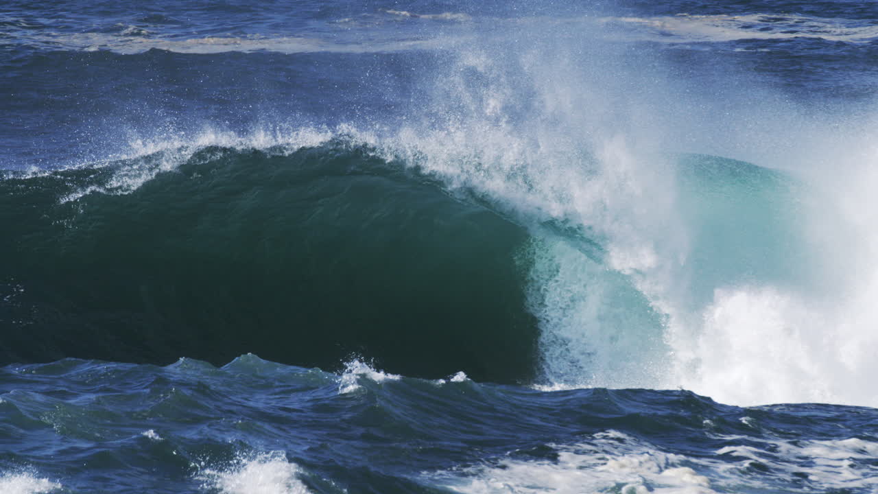 Super slow motion wave curls over, textured spray lifts from its crest in sunlight
