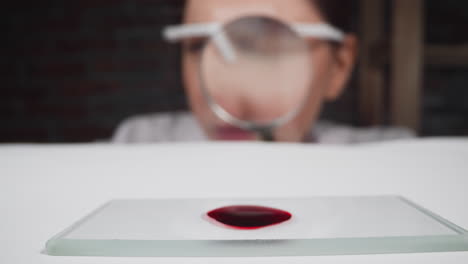 woman with magnifier glass looks at blood drop on table