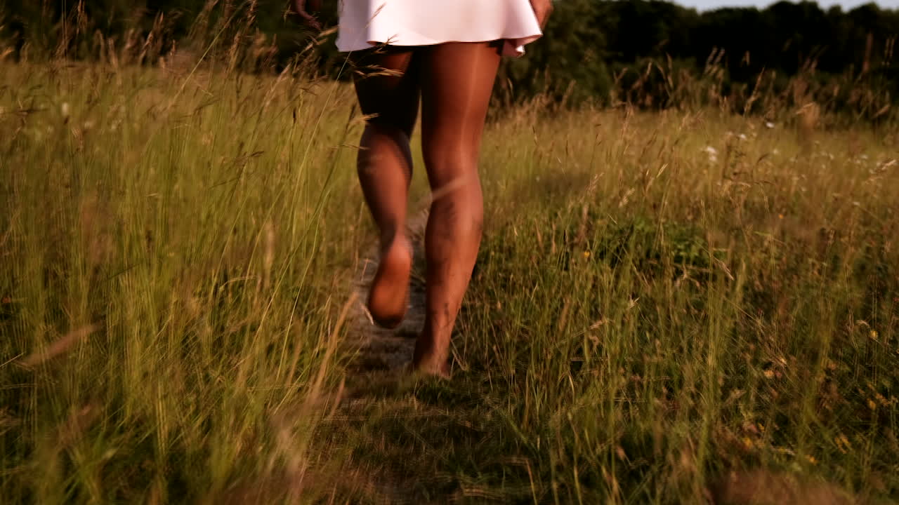 Black woman in skirt walks barefoot through path in field with high grass during a warm summer day