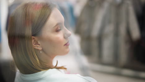 young woman with thoughtful expression seated indoors in mall environment, featuring soft lighting, subtle focus on hairstyle, delicate earrings, and blurred background with modern clothing displays