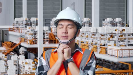 primer plano de ingeniero masculino asiático con casco de seguridad reza por algo mientras está de pie en el almacén con estantes llenos de mercancías de entrega