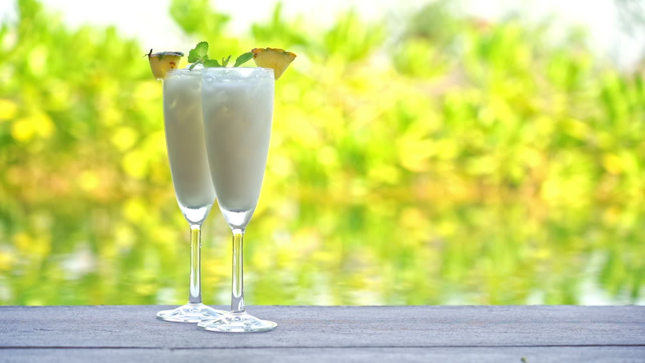 Two tropical drinks sit on a wooden table with water and green tropical plants in the background