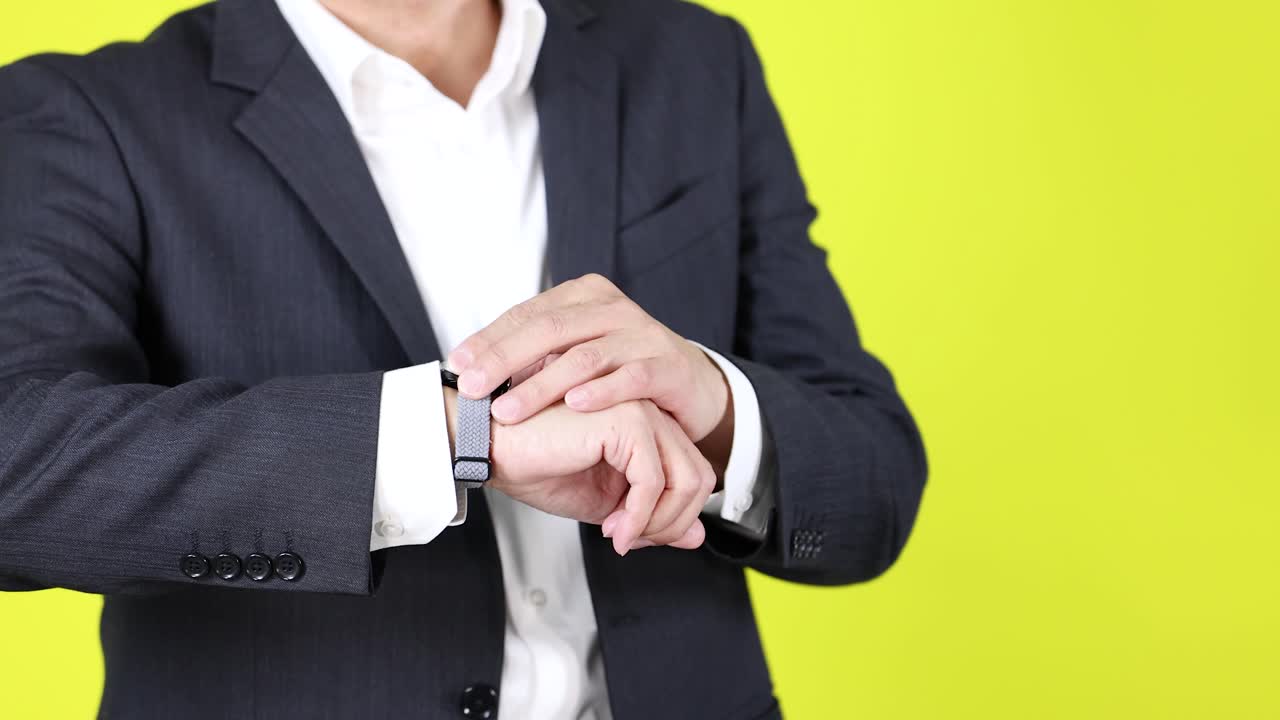 Man in suit urgently checks wristwatch, expressing stress, against bright yellow studio background