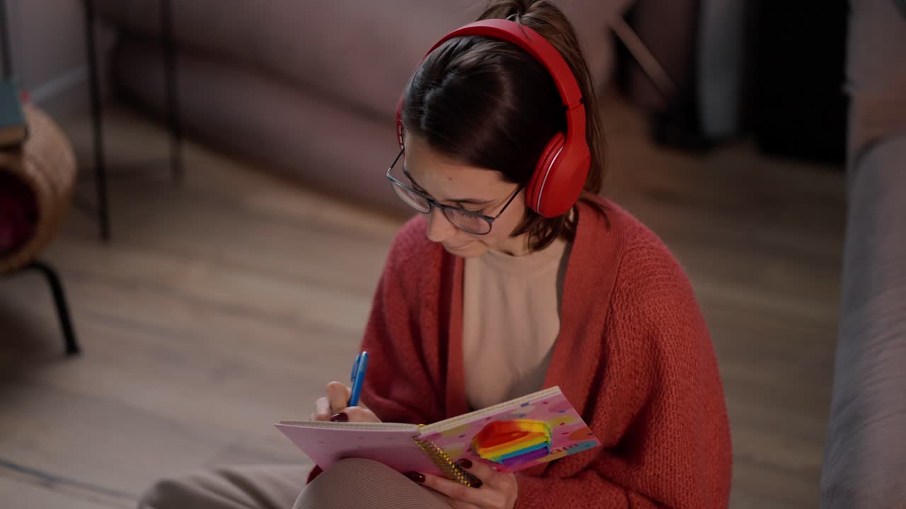 una confiada chica morena con auriculares inalámbricos rojos, gafas y un suéter rojo se sienta en el suelo de un apartamento moderno y hace notas en su cuaderno