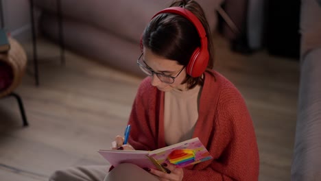una confiada chica morena con auriculares inalámbricos rojos, gafas y un suéter rojo se sienta en el suelo de un apartamento moderno y hace notas en su cuaderno