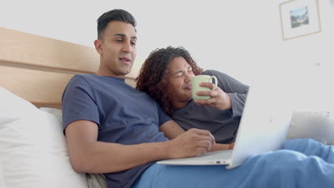 Happy-diverse-gay-male-couple-on-bed,-having-coffee-and-using-laptop-in-the-morning,-slow-motion