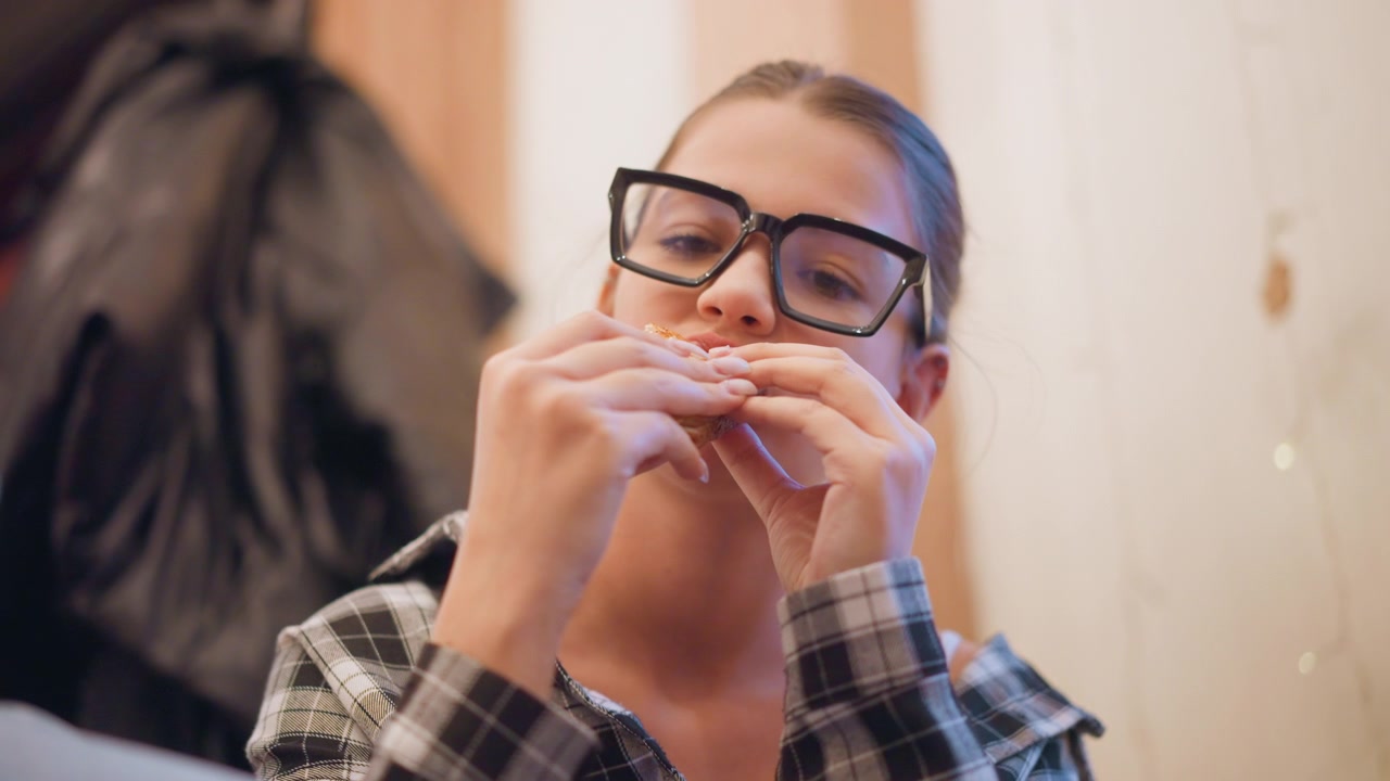 Close up of woman with glasses and plaid shirt eating food indoors. Woman holds item near her mouth focusing on consuming it while wearing eyeglasses. background includes soft lighting