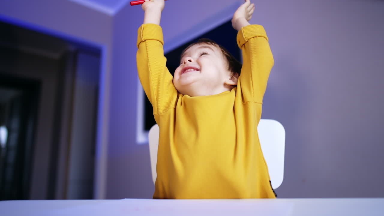 Baby boy drawing with red pen. Lovely toddler raises hands rejoicing cheerfully. Close up.