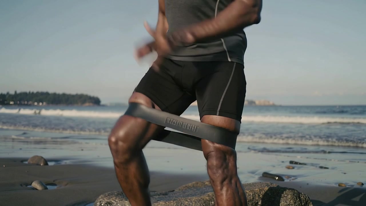 Homem de bermuda de compressão dando passos laterais com elástico em uma praia rochosa após o ajuste do elástico, durante o treino