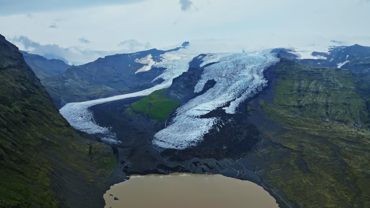 Dronebillede af en islandsk gletsjer, der flyder ud i en mudret proglacial sø, omgivet af stejle, grønne bjerge og mørk vulkansk moræne under en skyet himmel.