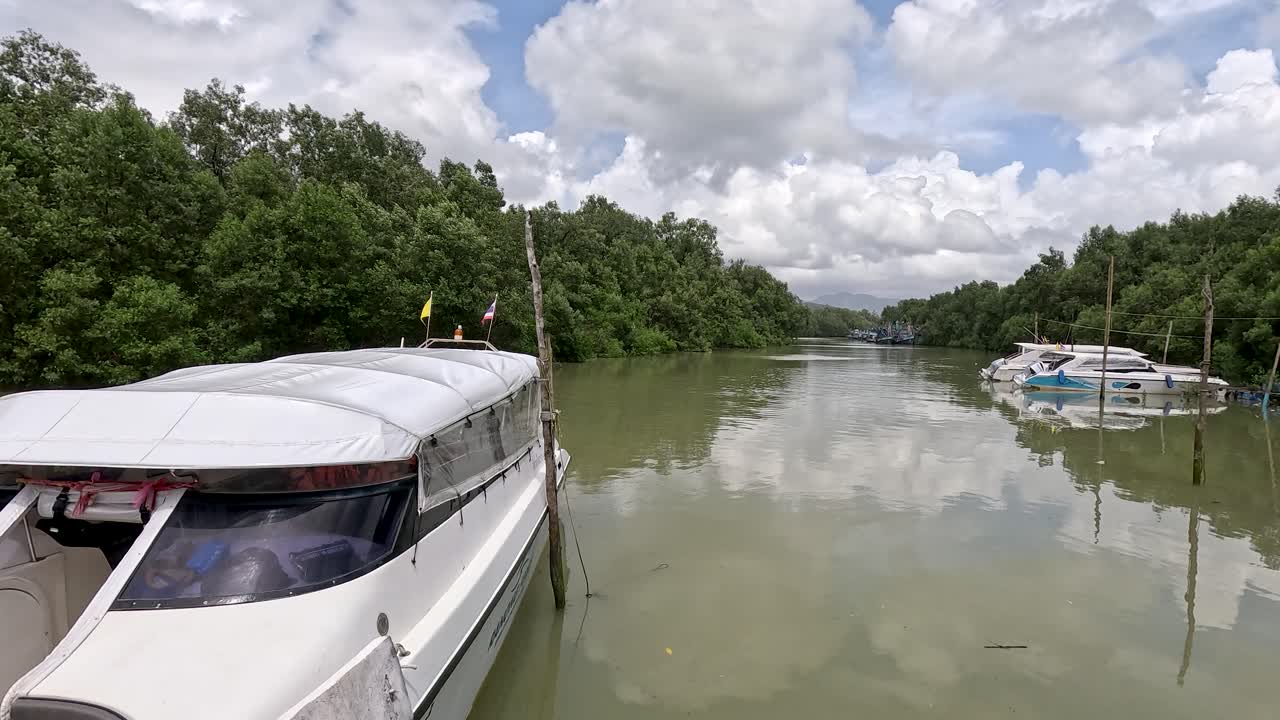 A stationary speedboat is docked at a calm mangrove river pier under bright daylight, with the camera slowly panning to reveal more of the lush riverside and additional boats