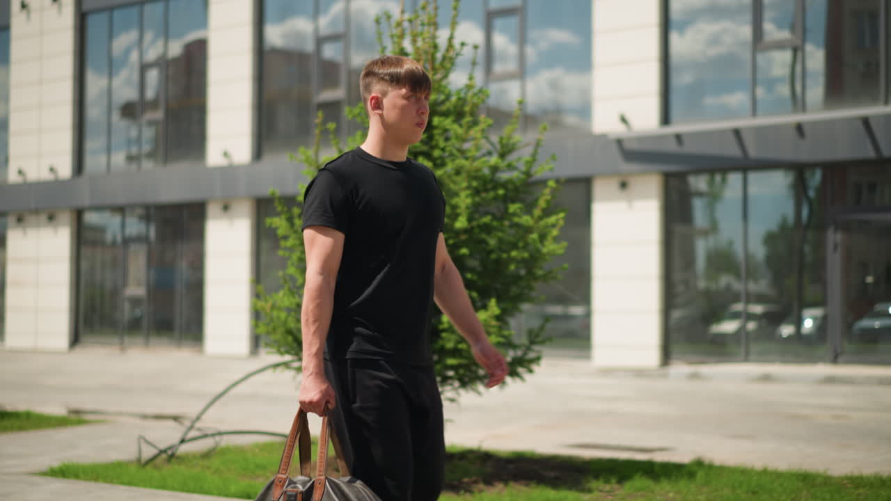 흰색 양복을 입은 남자가 가죽 더플백을 들고 유리 사무실 건물을 자신감 있게 걸어가고 있습니다. 현대적인 건축물과 반사되는 창문이 전문적인 도시 풍경을 연출하며, 맑은 날씨가 선명도를 높입니다.