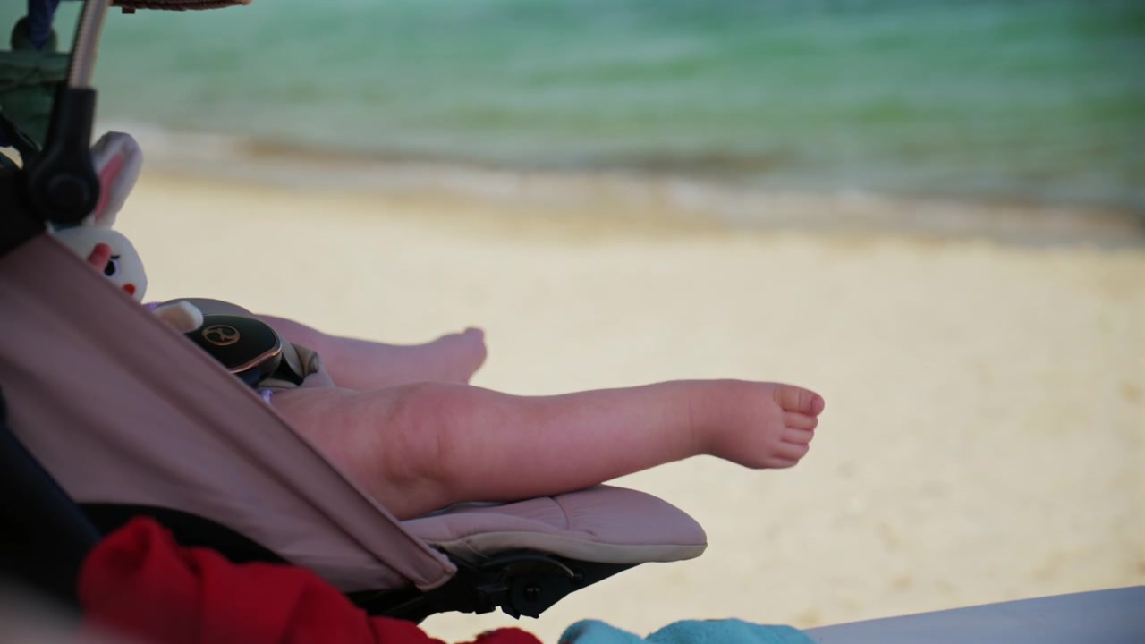Strandscene med blødt fokus, hvor en babys fødder hviler i en klapvogn, med sandstrand og turkisblåt hav sløret i baggrunden.