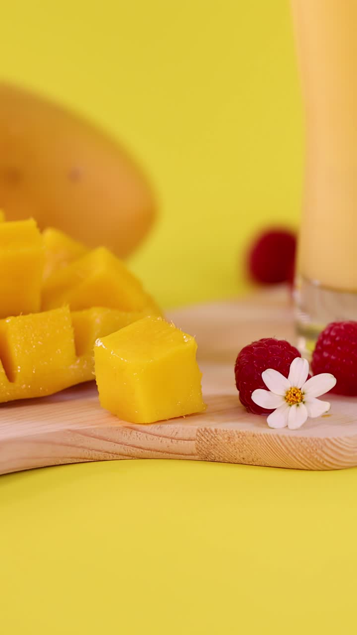 Cubetti di mango, smoothie, lamponi e fiori disposti su un tagliere con illuminazione brillante.