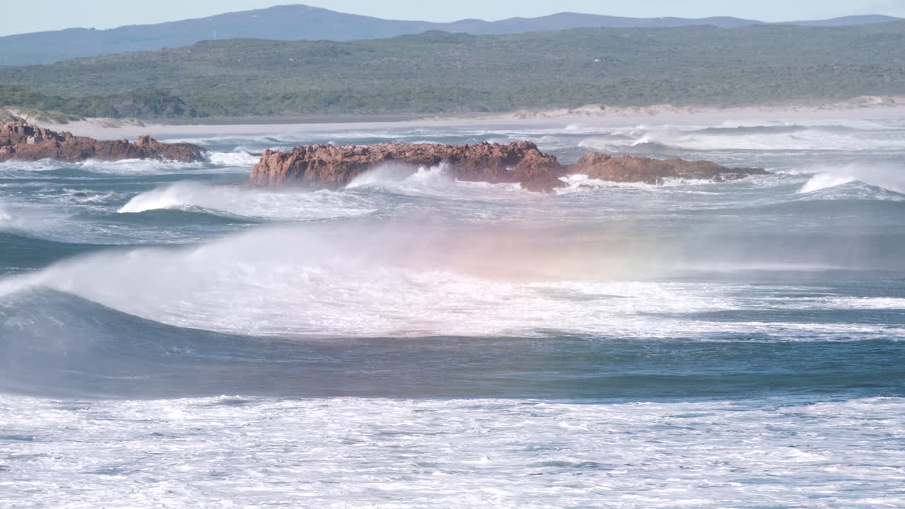 Tumultuous ocean waves roll in toward coastline, wind blows sea spray over crest