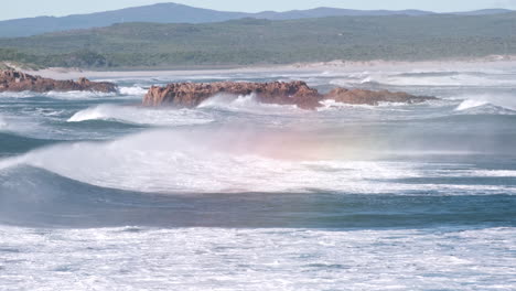 tumultuous ocean waves roll in toward coastline, wind blows sea spray over crest