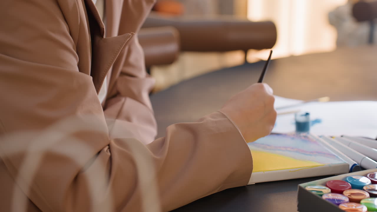 Manos mezclando la paleta de acuarelas en el escritorio, un pincel fino mojándose en los pocillos de pigmento, resplandor de la lámpara y reflejo del aro de luz, pinceladas deliberadas y técnica firme durante la sesión de arte de mesa