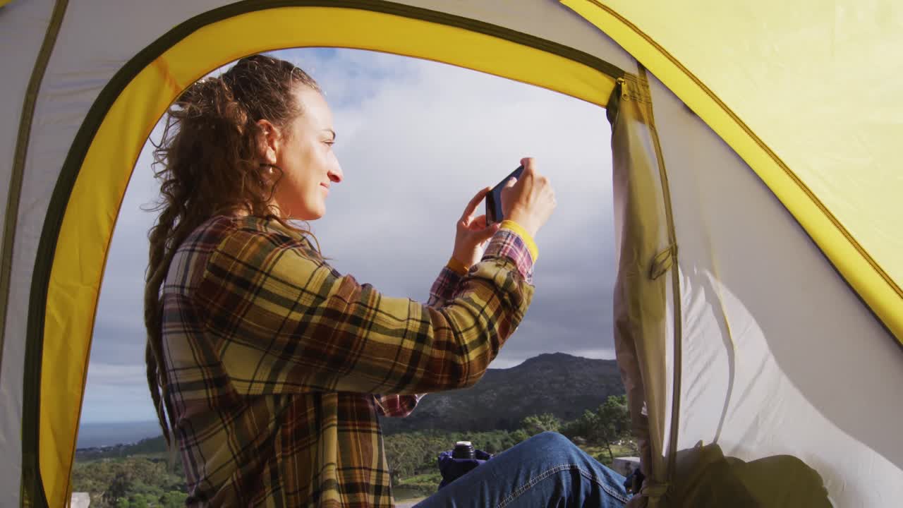 feliz mujer caucásica acampando, sentada fuera de la tienda en la montaña rural tomando fotos con el teléfono inteligente