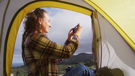 happy caucasian woman camping, sitting outside tent on rural mountain taking photo with smartphone