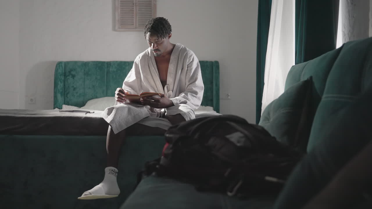Man in bathrobe sitting on bed reading book, soft light streaming through curtains. Relaxed morning scene, wearing socks and sandals, backpack on bed. Calm, peaceful atmosphere