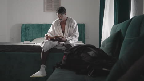 man in bathrobe sitting on bed reading book, soft light streaming through curtains. relaxed morning scene, wearing socks and sandals, backpack on bed. calm, peaceful atmosphere