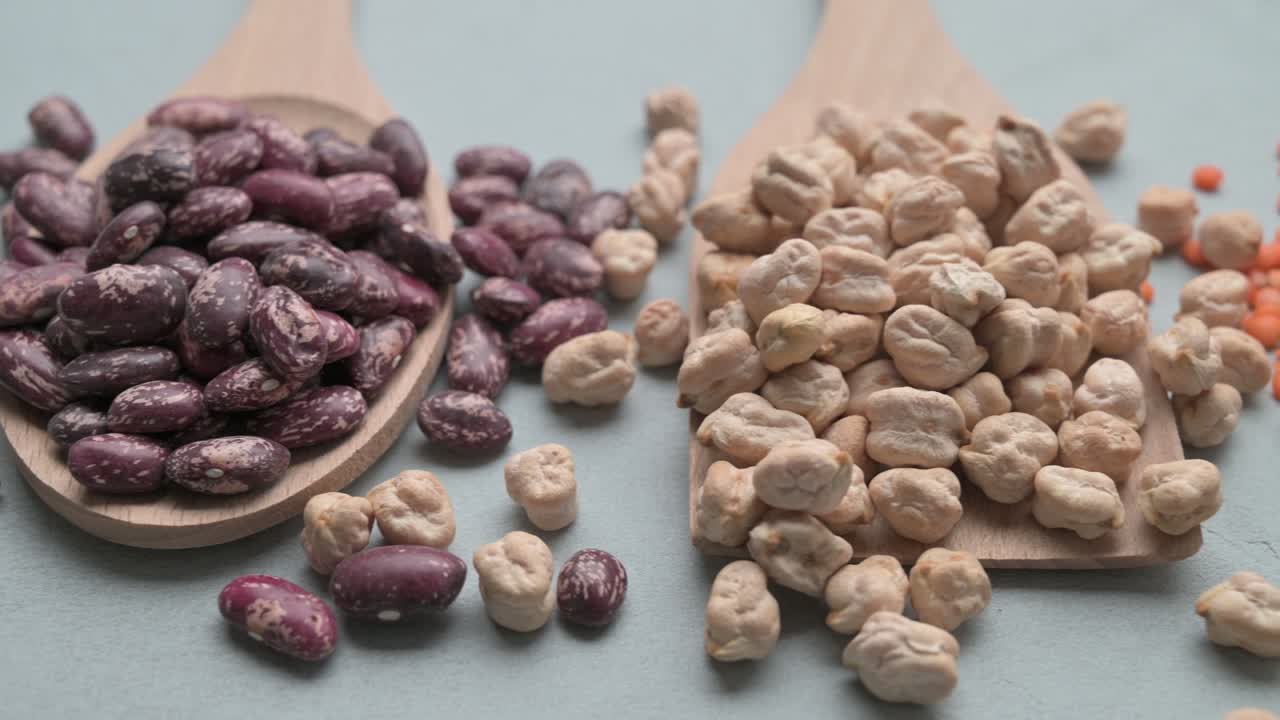 Different types of beans in wooden spoons on gray stone table.
