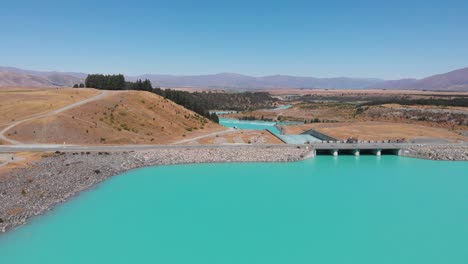 beautiful aerial view of the turquoise water of beautiful pukaki lake on sunny summer day in 4k with a car driving through the narrow bridge and dam