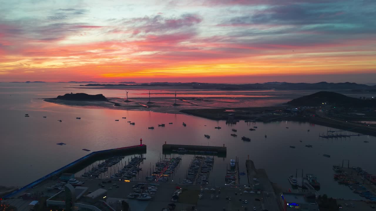Baglæns afslørende luftfoto af silhuetter af Jeongok Marina og bytage, Nue Island Tando havns vindmøller mod en flammende skyformation efter solnedgang med spredte fiskerbåde over en rolig tidevandsbugt.