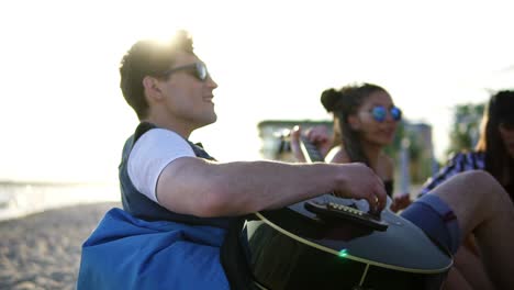 Young-man-playing-guitar-among-group-of-friends-sitting-on-easychairs-on-the-beach-and-singing-on-a-summer-evening-during-a-sunset.-Slowmotion-shot
