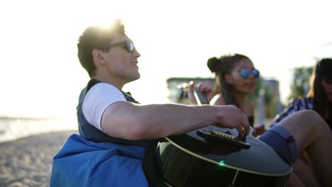 Junger-Mann-Spielt-Gitarre-In-Einer-Gruppe-Von-Freunden,-Sitzt-Auf-Sesseln-Am-Strand-Und-Singt-An-Einem-Sommerabend-Bei-Sonnenuntergang.-Zeitlupenaufnahme