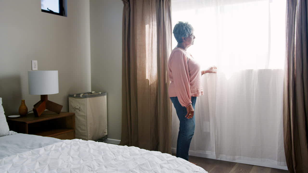 Donna anziana che soffre di depressione in piedi vicino alla finestra della camera da letto a casa