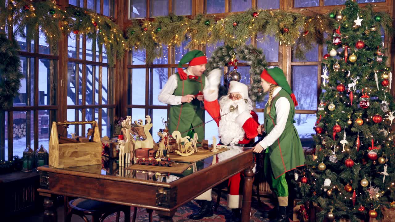 Santa Claus is sitting in specially decorated room at the table, holding the telephone. New year treeand high windows in the background. Christmas spirit concept close up video. Christmas eve. New Year is coming. Two elves near Santa Claus are listening to instructions.