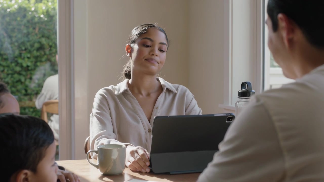 Scrolling woman in light beige button-up navigating tablet at kitchen table, prompted by children