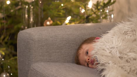 baby boy falling asleep on living room sofa on christmas night. decorated tree in the background