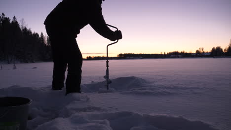 man drills through snowy ice to prepare for ice fishing at dusk