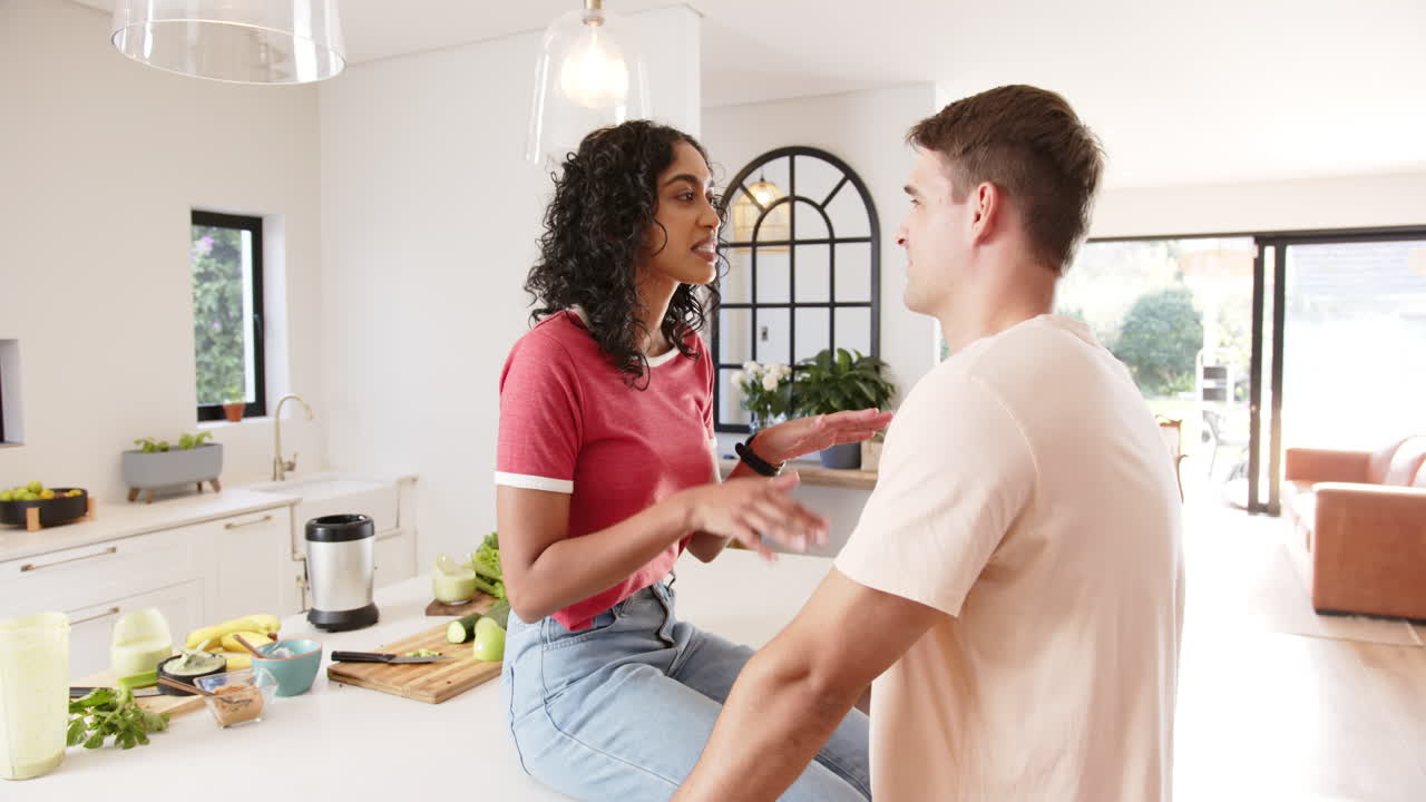 Young multiracial couple sitting on kitchen counter, holding hands and talking lovingly, at home