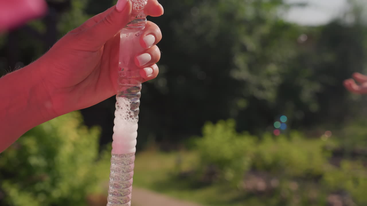 CloseUp White Woman Squeezing Textured Tube, Pink Foam Crystals And Gel Bubbles Glint In Sunlight Over Green Garden Background, Manicured Nails Press And Twist For Satisfying Sensory Stress Relief