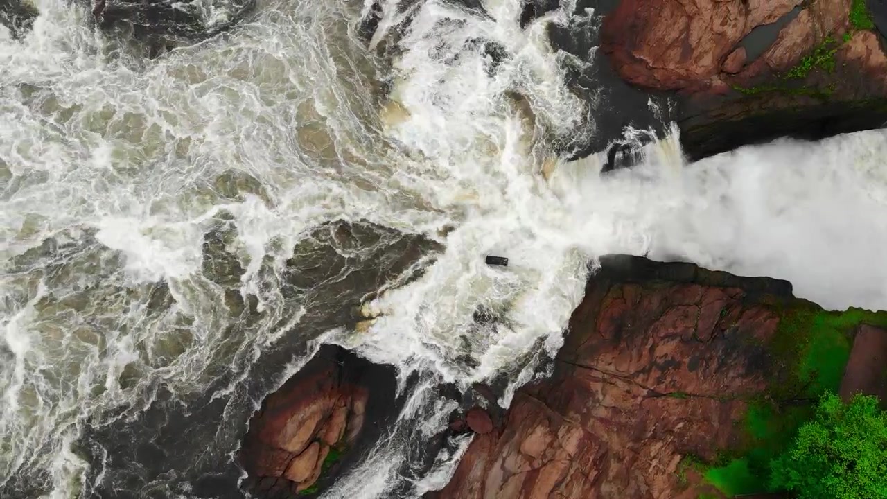 Top-down aerial view of powerful whitewater rushing over dark rocks and red stone with green vegetation along the edge in bright daylight.