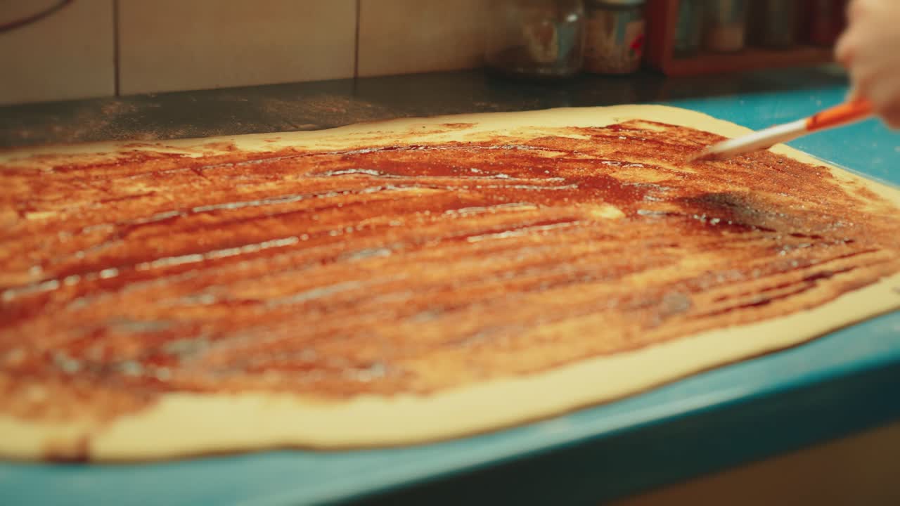 Primo piano di mani adolescenti che usano una spatola per stendere un impasto di cannella, zucchero e burro sulla pasta stesa. Ricca consistenza del ripieno dolce per i tradizionali cinnamon rolls