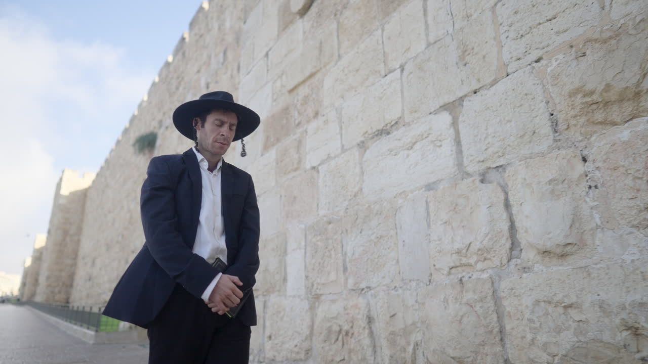 전통적인 옷을 입은 한 남자가 예루살렘의 고대 석벽에 엄숙하게 서서 깊은 성찰과 문화적 존경의 순간을 포착합니다.