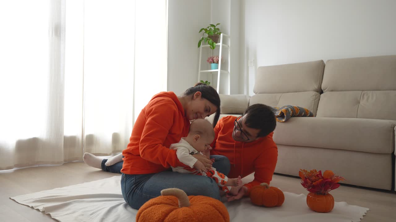 Family time of a father playing and talking to his 4 months old baby sitting on his mum's knees over a blanket surrounded by pumpkins in the living room