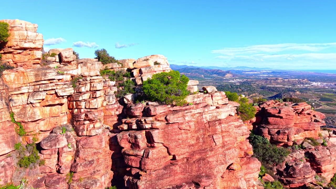 Acantilado rocoso con vistas a la llanura costera y al mar Mediterráneo, España. Vista lateral de una cornisa de arenisca roja con un árbol verde, campos de cultivo en mosaico, ciudad costera y horizonte azul del océano bajo un cielo despejado.