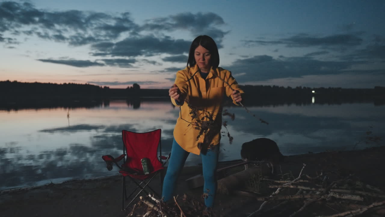 Al tramonto, una donna in campeggio in riva al lago accende un falò con un fiammifero e accendifuoco, accanto a un lago calmo e riflettente, con rami e una sedia pieghevole nelle vicinanze. Scintille e un bagliore caldo contrastano la frescura della sera, creando un momento tranquillo e gioioso.