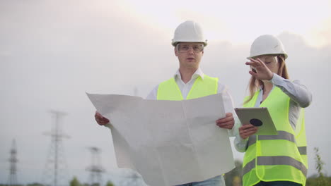 Ingeniería-Trabajando-En-Una-Torre-De-Alto-Voltaje-Verifique-La-Información-En-La-Tableta-Dos-Empleados,-Hombre-Y-Mujer