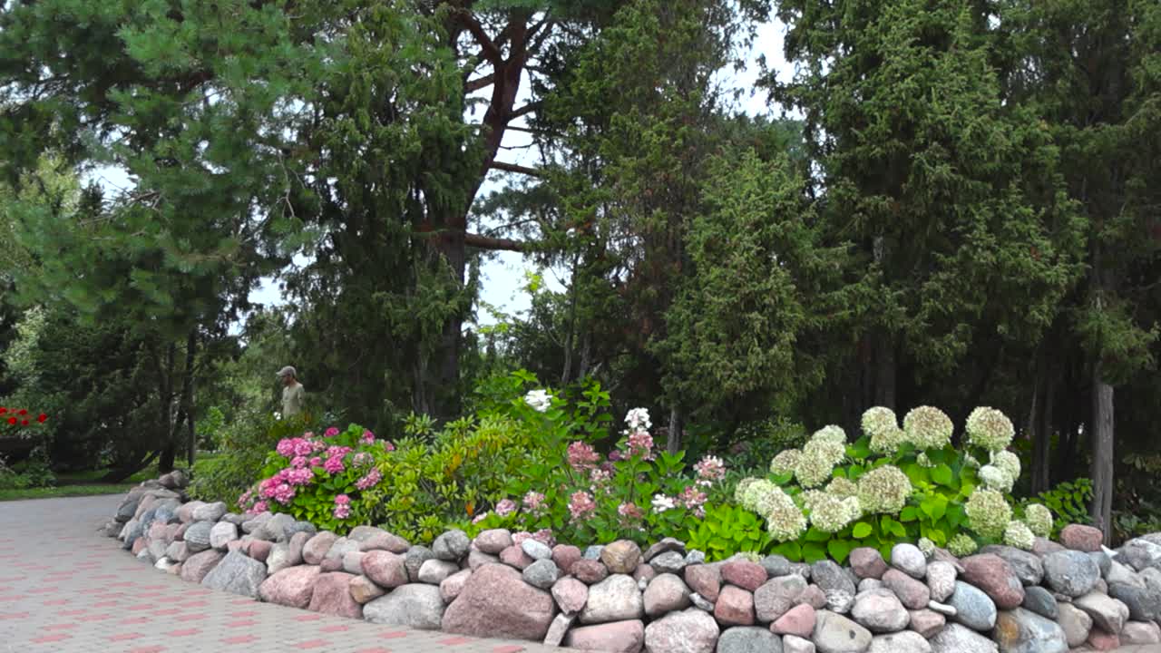 Flower bed filled with white, pink and colorful flowers and plants that's surrounded by beach rocks forming a beautiful garden during summer time. A road surrounds the garden and trees in the back.